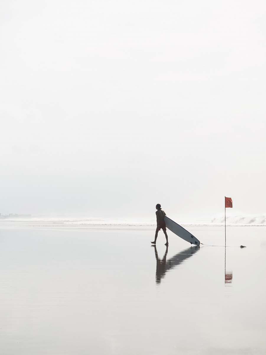 double six beach man walking while dragging surfboard at the beach near flag during day indonesia